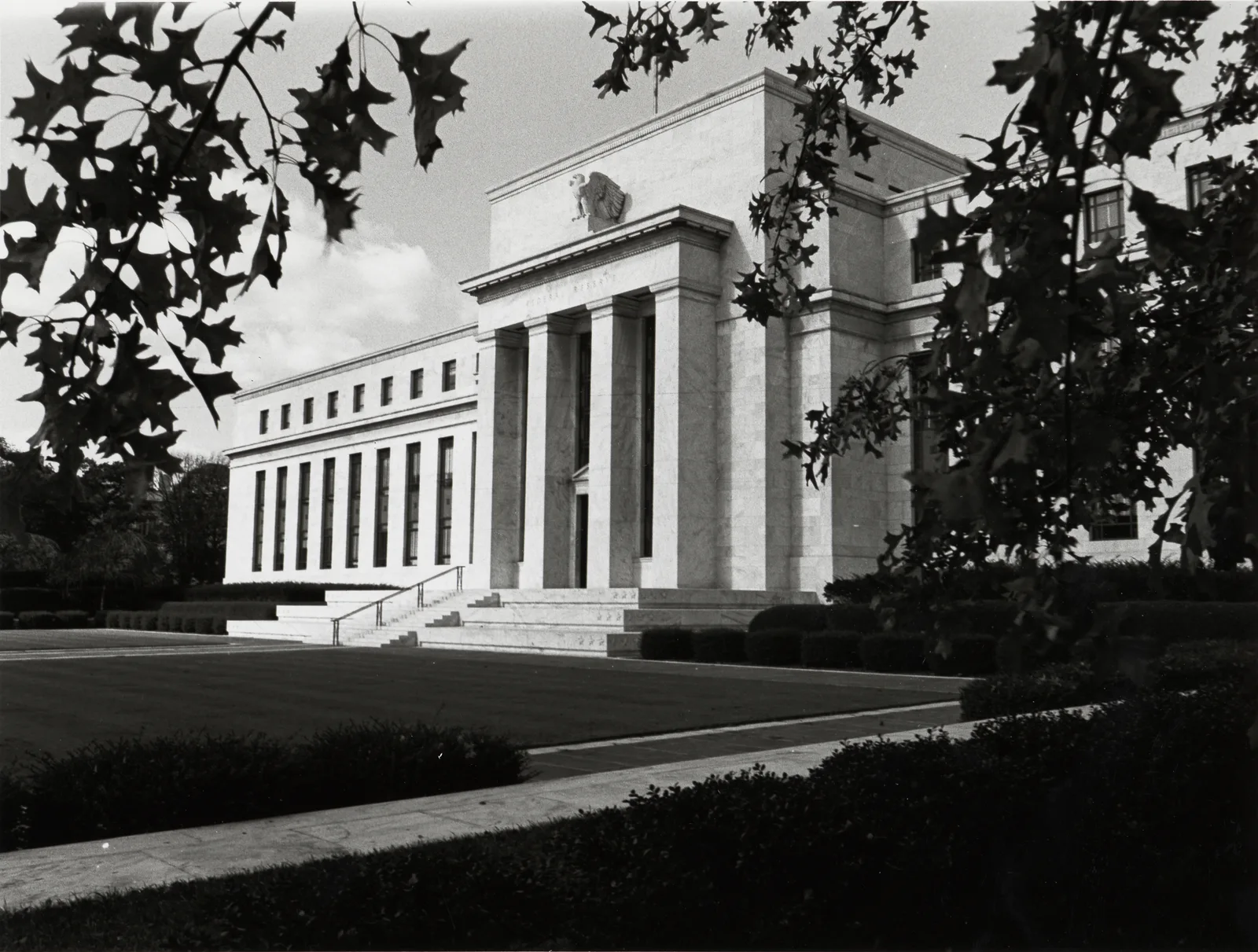 The Marriner S. Eccles Federal Reserve Board Building in Washington, D.C., a marble Beaux-Arts style building completed in 1937, framed by tree branches with an eagle sculpture above its columned entrance.