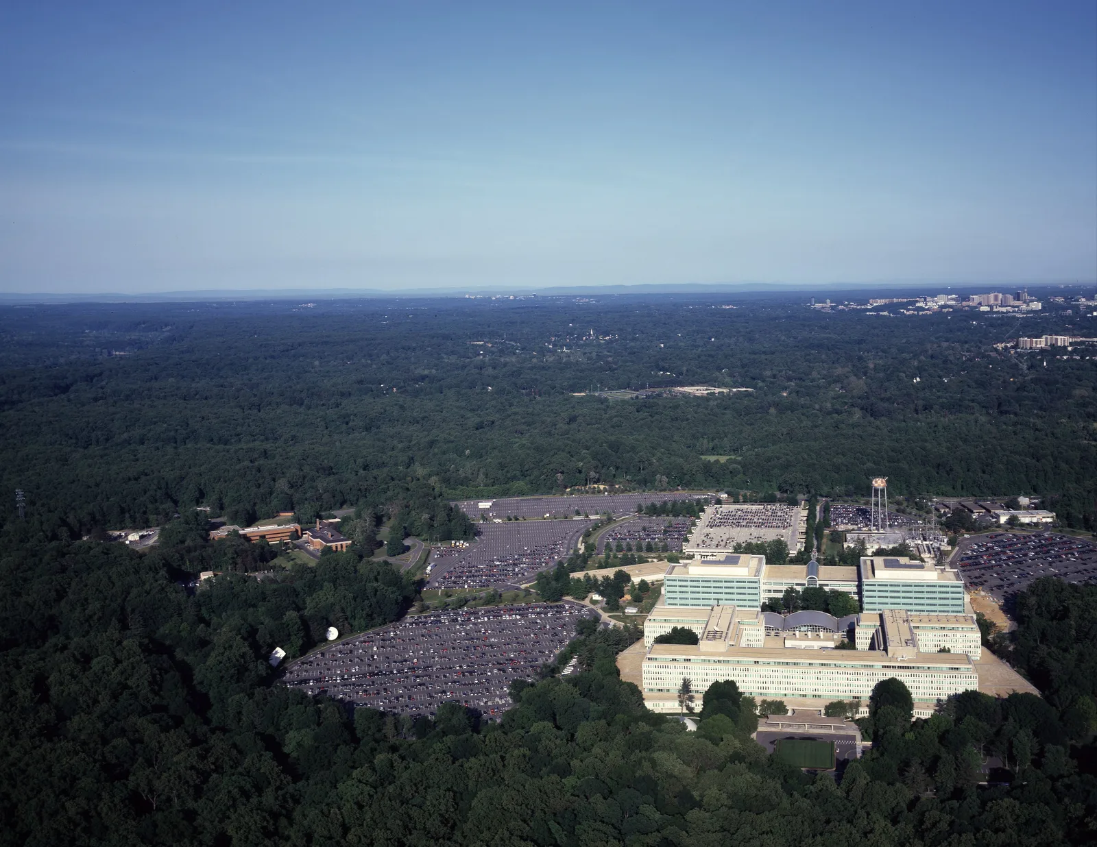 Aerial view of CIA headquarters in Langley, Virginia: white office buildings and wide parking lots in a clearing surrounded by dense green woods, with the Washington, D.C., area on the horizon