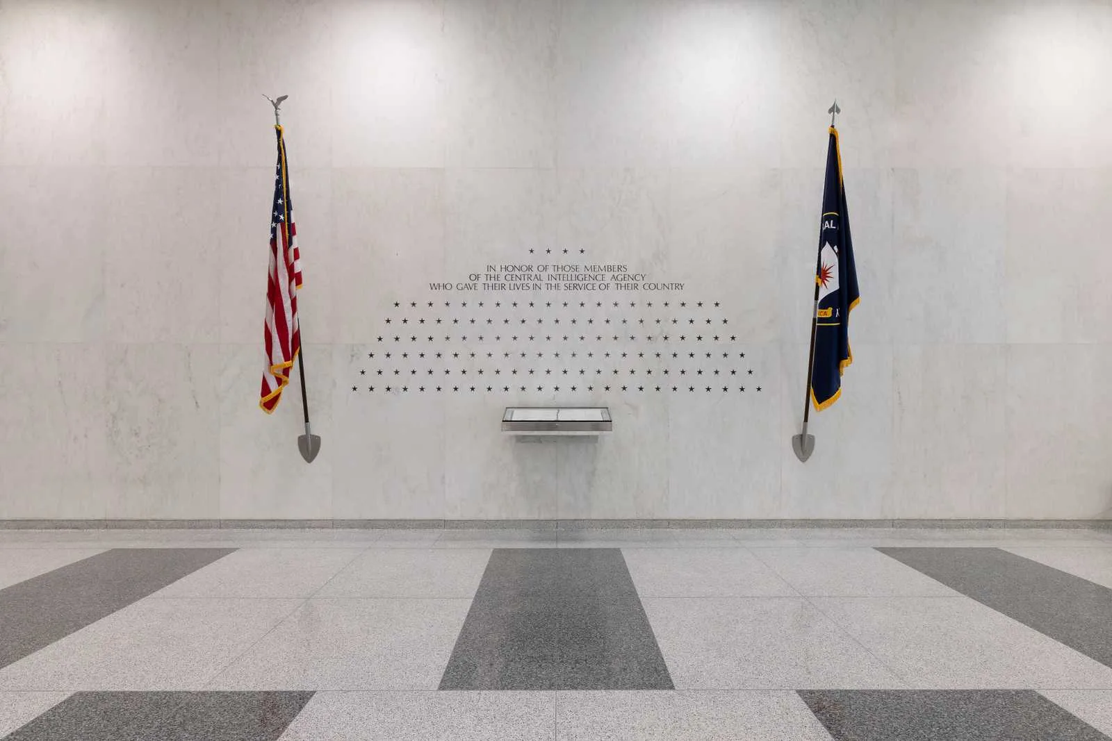 The CIA Memorial Wall at agency headquarters: rows of carved stars on a white marble wall above a small glass case holding the Book of Honor, between the American flag and the CIA flag