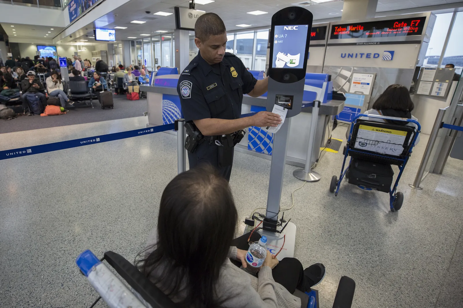 A U.S. Customs and Border Protection officer operates an NEC biometric facial-recognition kiosk at a United Airlines international departure gate in Houston, processing a seated traveler whose face is photographed and matched against government records before boarding.