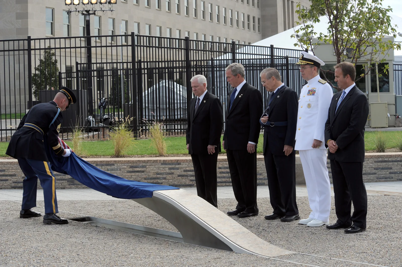 Officials stand beside the first inscribed bench unveiled at the Pentagon Memorial dedication, with rows of cantilevered memorial benches across a gravel field.