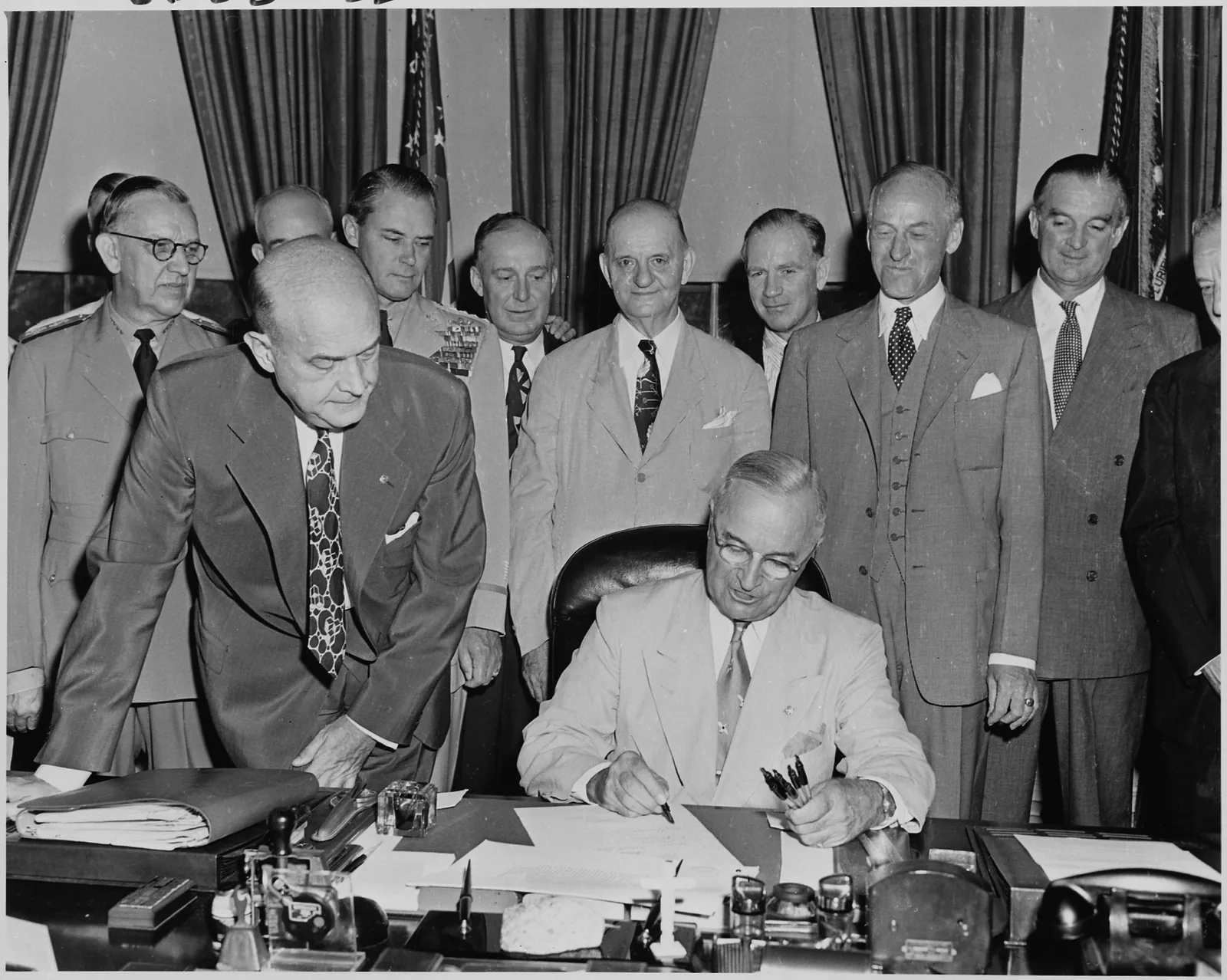 President Harry Truman seated at his desk in the Oval Office signing the National Security Act Amendments of 1949, with officials gathered around the desk.
