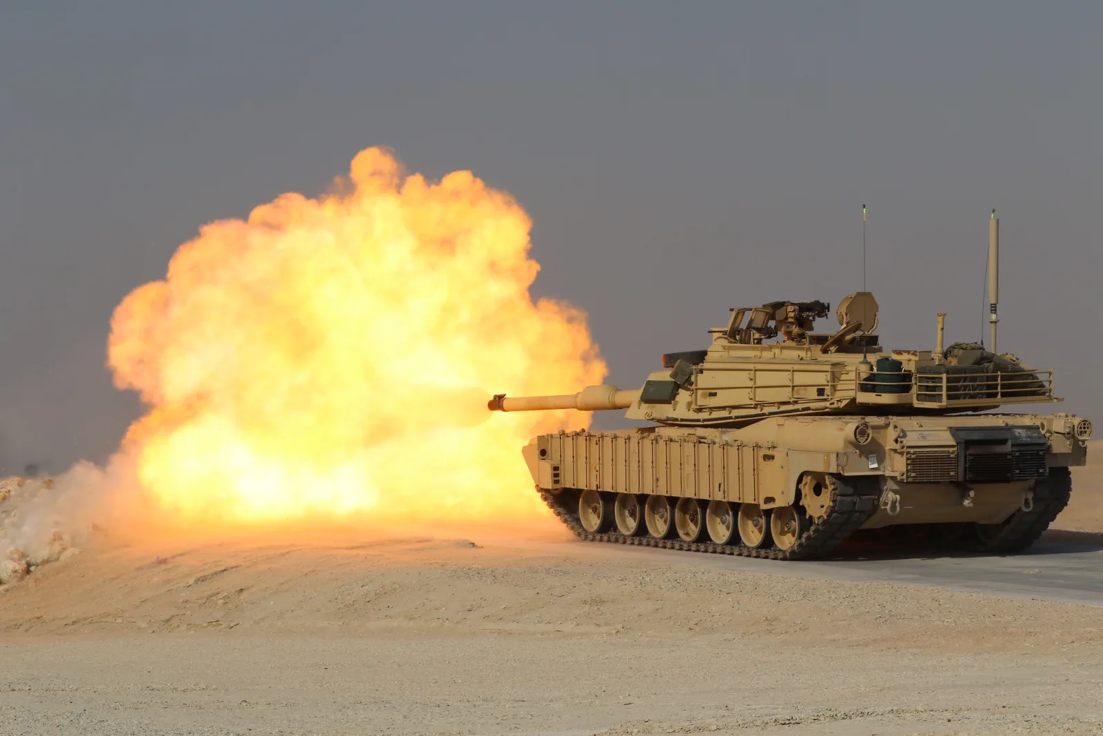 A U.S. Army M1A2 Abrams tank firing its main gun on a training range, with a ball of fire and smoke erupting from the muzzle