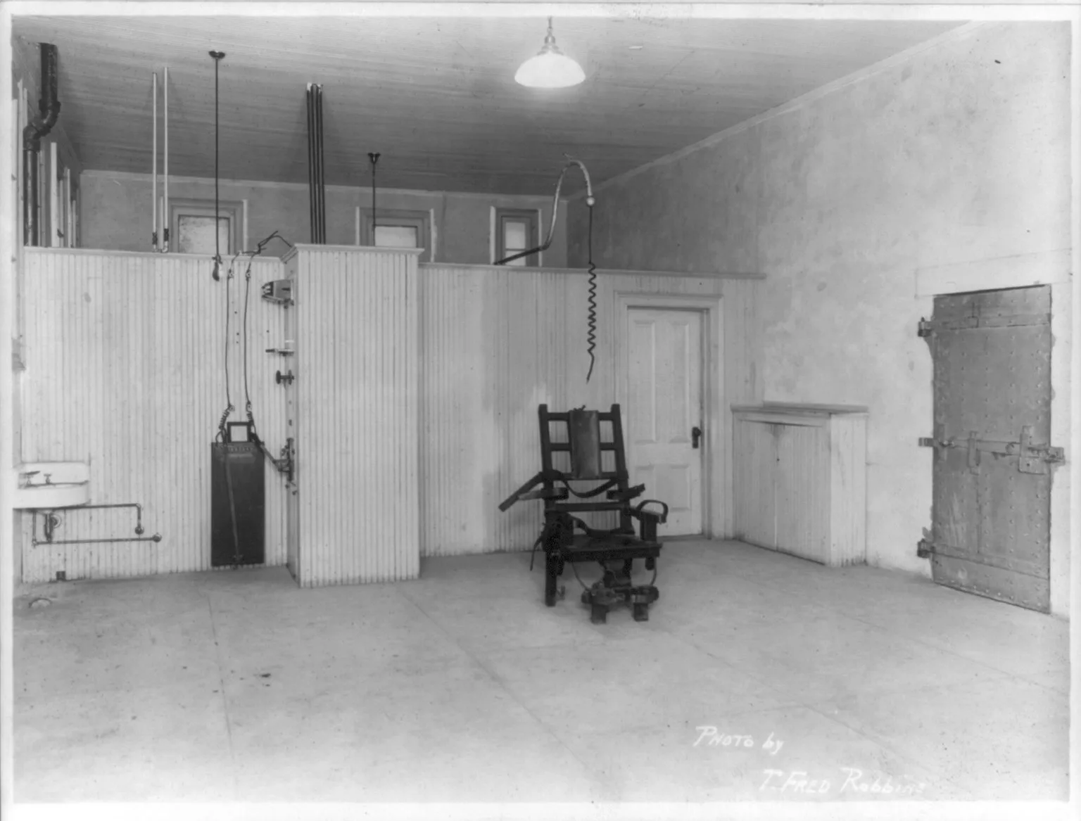 The electrocution chamber at Sing Sing Prison in New York in 1915: a bare room with a heavy wooden electric chair fitted with leather straps, an electrode cable hanging from the ceiling above it, wood-paneled walls, and a riveted steel door
