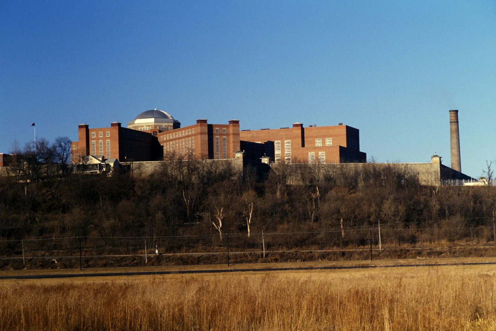 The old U.S. Disciplinary Barracks at Fort Leavenworth, Kansas, in 1977: tall red brick cell blocks topped by a domed rotunda, a stone perimeter wall with guard towers, and a smokestack, seen across a winter field