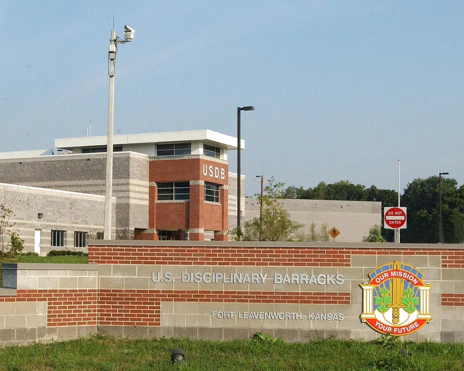 The entrance sign of the current U.S. Disciplinary Barracks at Fort Leavenworth, Kansas, in front of a modern brick and concrete building marked U.S.D.B., with a security camera mast