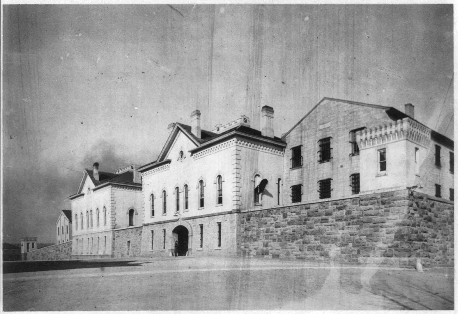 An early 1900s photograph of the U.S. Military Prison at Fort Leavenworth: long stone and brick buildings with barred windows above a heavy masonry wall and an arched gateway