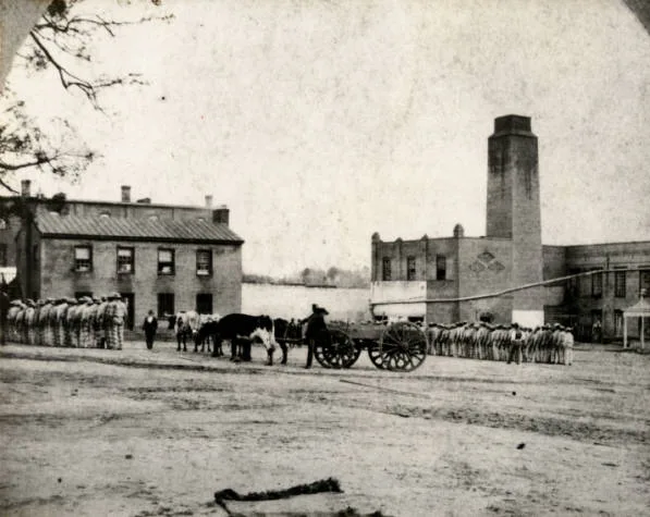 The yard of the Texas State Penitentiary at Huntsville in the 1870s: long lines of prisoners in striped uniforms stand beside an ox-drawn cart, in front of brick prison buildings and a tall chimney. The same prison, now the Huntsville Unit, is where Texas carries out executions.
